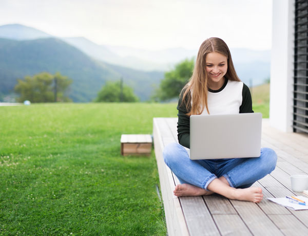 A young woman sits on a porch with her laptop in a rural setting.